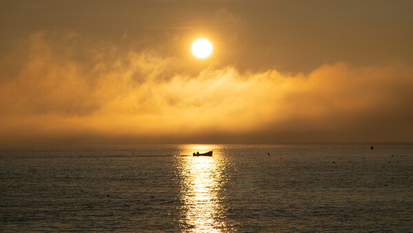 Fishing boat silhouetted against golden sunlight reflecting on calm ocean water at sunset.