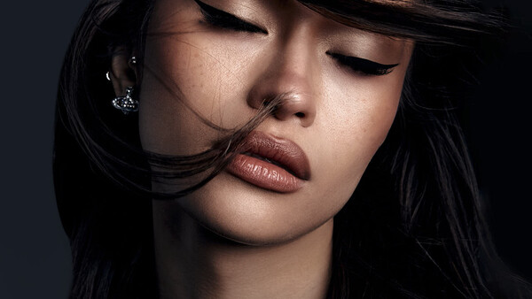 Woman with long black hair and closed eyes, wearing a chain necklace against a dark background.