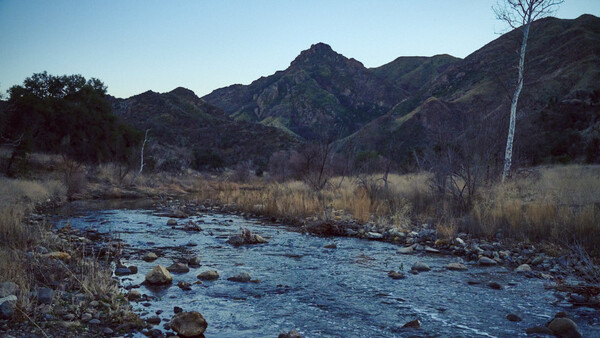 Mountain stream with rocks flowing through a valley with sparse winter vegetation and distant peaks.