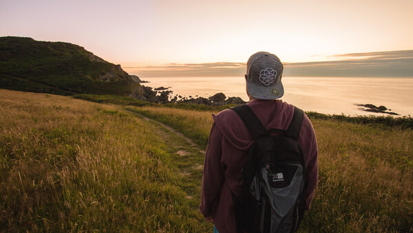 Hiker in maroon jacket and cap with backpack standing on grassy hillside at golden hour overlooking calm water