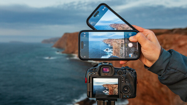 Hand holding smartphone and DSLR camera both displaying the same coastal cliff landscape with ocean.