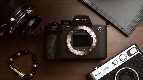 Sony a7 mirrorless camera body with interchangeable lenses and vintage film camera arranged on brown surface.