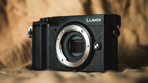 Panasonic Lumix mirrorless camera body with lens mount exposed, positioned on sandy surface with warm-toned background.