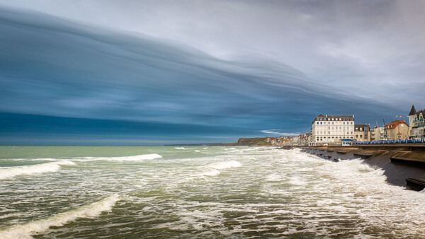 Dramatic coastal town with white-capped waves and moody sky reflected in water.