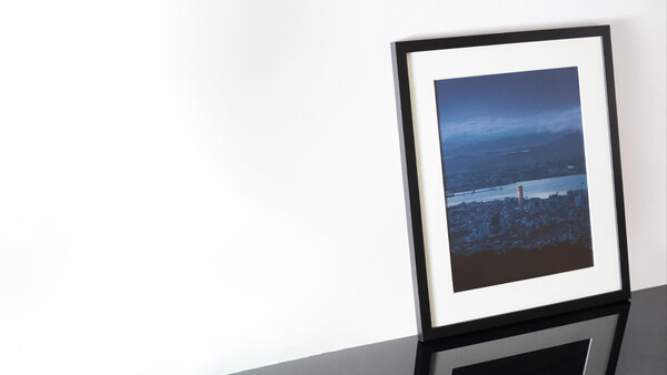 Framed photograph of a nighttime cityscape with illuminated buildings and water reflection displayed on a white wall.
