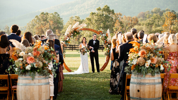 Outdoor wedding ceremony in a vineyard valley with a wooden arch and flower arrangements flanking the aisle.