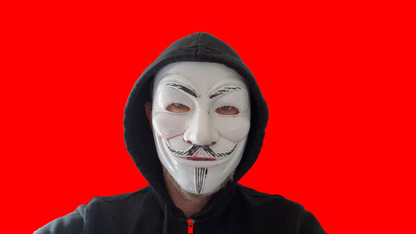 Person wearing a Guy Fawkes mask and black hoodie against a bright red background.