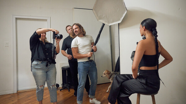 Photography studio setup with model posing on stool while crew adjusts octabox softbox lighting.