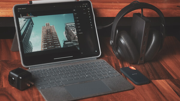 Tablet displaying architectural photography with editing software open, surrounded by headphones and external hard drive on wooden surface.