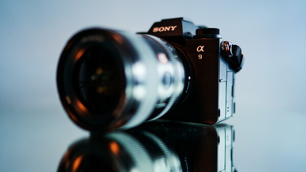 Sony α9 mirrorless camera with telephoto lens mounted, photographed against a neutral sky background with reflective surface below.