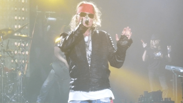 Performer on stage wearing sunglasses and leather jacket, singing into microphone with dramatic yellow stage lighting.