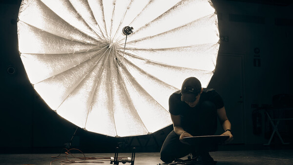 Photographer crouching beside a large parabolic reflector on a dark studio floor.
