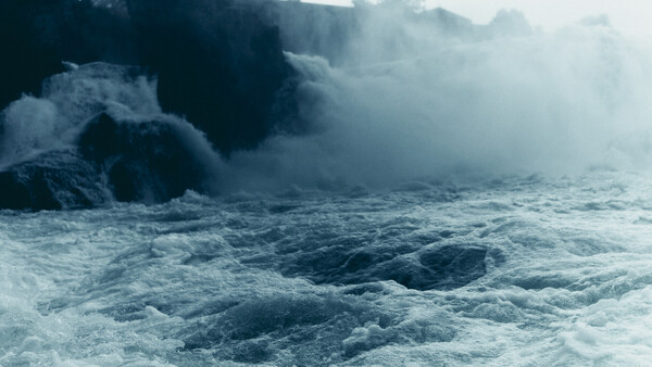 Powerful waves crash against dark rocky cliffs shrouded in white sea spray and mist.