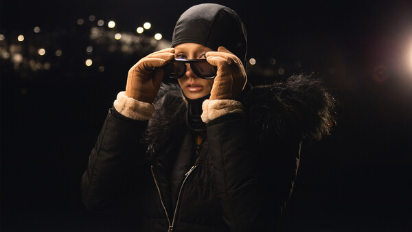 Person wearing dark winter jacket and sunglasses adjusting glasses against dark background with warm bokeh lights.