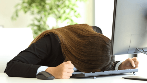 Woman in black blazer slumped over desk with head down, hand on keyboard, appearing exhausted or overwhelmed.