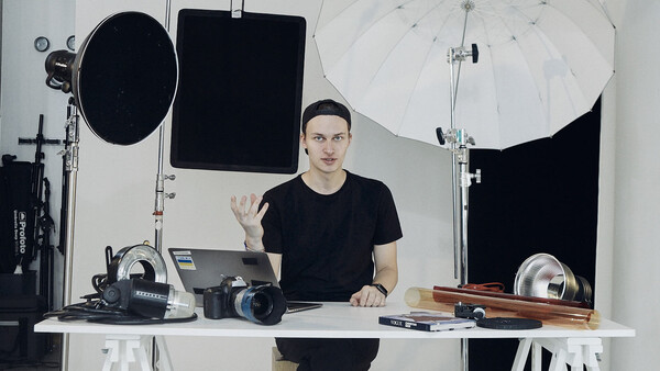 Photographer gesturing while seated at desk surrounded by professional camera equipment and lighting gear.
