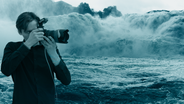 Photographer holding a DSLR camera while standing before massive ocean waves crashing with white spray.