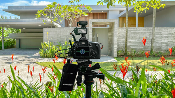 Professional video camera on tripod filming a modern residential house surrounded by tropical plants.