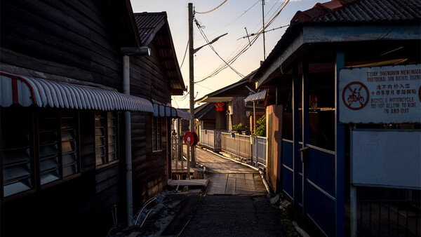Narrow alley lined with dark wooden buildings at dusk, with warm golden light illuminating the street and distant figures walking.