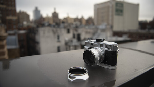 Vintage rangefinder camera with lens cap displayed on a rooftop overlooking an urban cityscape.