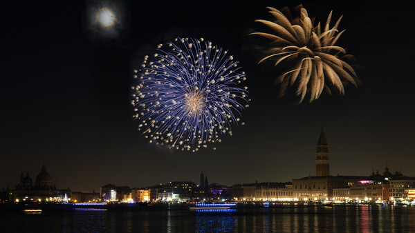 Blue and gold fireworks exploding over a waterfront cityscape at night with illuminated buildings reflected in calm water.