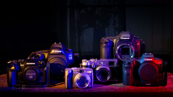 Collection of Canon and Olympus digital cameras illuminated with blue and red LED lighting against a dark background.