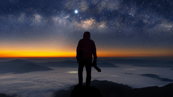 Silhouetted photographer standing on a mountain peak at twilight, facing a starry sky with the Milky Way visible above an orange and gold sunset horizon.