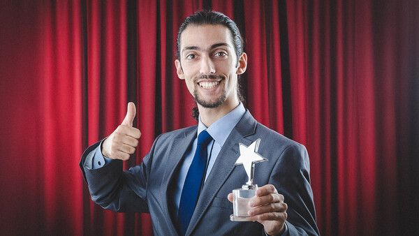 Professional man in business suit holding star-shaped award with thumbs up gesture against red curtain backdrop.