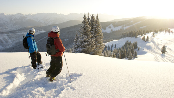 Two hikers in winter gear walking through deep snow on a mountain slope with evergreen trees and snow-covered valleys visible below.