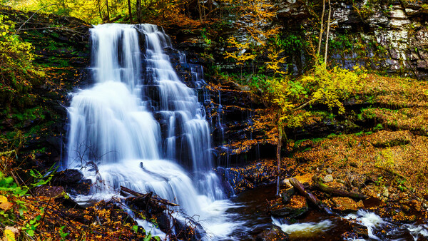 Cascading waterfall flows over rocky ledges surrounded by autumn foliage and moss-covered stone.