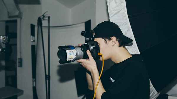 Photographer in black cap and shirt holding a professional video camera with yellow shoulder strap in a studio setting.