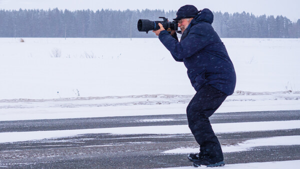 Photographer in dark winter coat crouched on frozen lake while shooting with telephoto lens, forest treeline visible across snowy landscape.