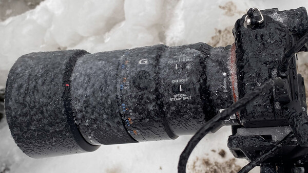 Telephoto lens covered in frost and ice mounted on camera body in snowy conditions.
