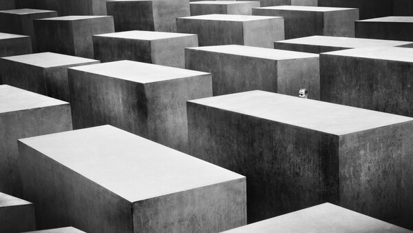 A small figure standing alone among towering concrete memorial blocks in a geometric maze-like formation.