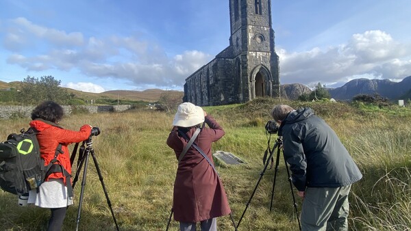 Three photographers with tripod-mounted telephoto lenses facing a stone church ruin in a mountain valley.