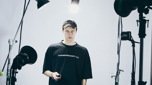 Man in black t-shirt and cap stands in professional studio surrounded by lighting equipment.