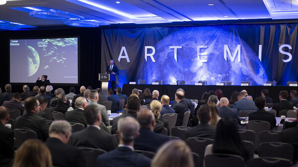 Speaker presenting at a conference with large blue screen displaying ARTEMIS backdrop.