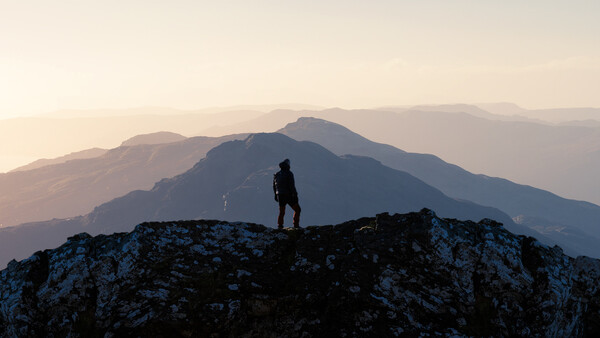 Silhouetted hiker standing on a rocky outcrop overlooking layered mountain ranges at sunset.