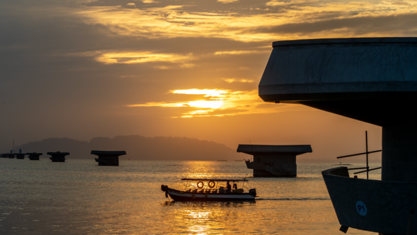 Silhouetted boats anchored in calm water at golden hour sunset with mountains in distant background.