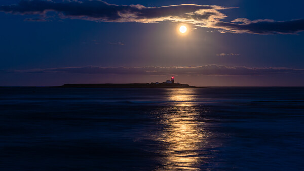 Full moon illuminating a golden path across calm ocean water toward a distant lighthouse at dusk.