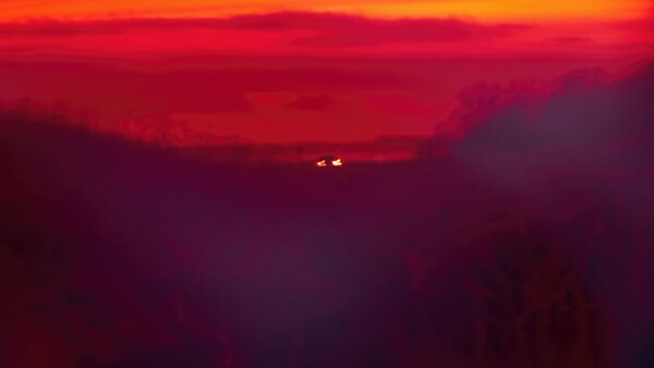 Silhouetted mountain ridges against a deep red and orange sunset sky with two distant lights visible on the horizon.