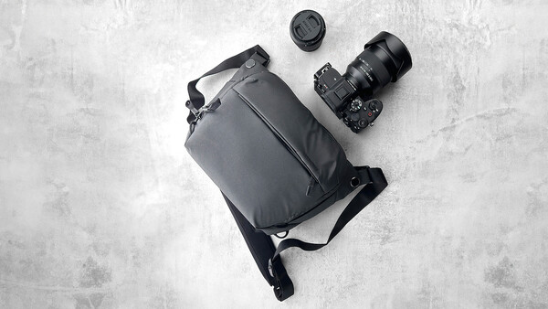 Flat lay of camera gear including DSLR body with attached lens, shoulder bag, and lens cap on concrete surface.