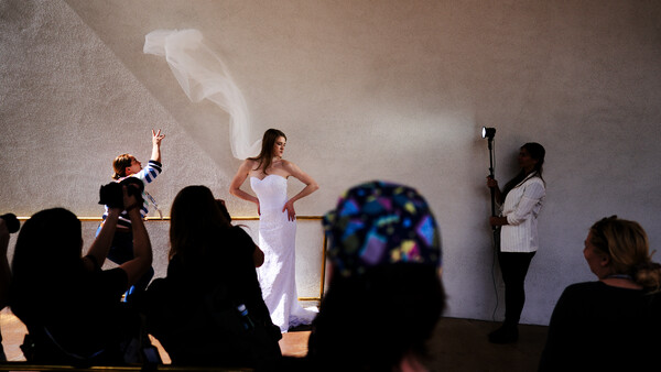 Woman in white dress posing on stage during a fashion or bridal show while photographers and spectators watch from the audience.