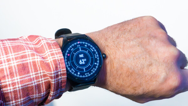 Smartwatch with blue digital display showing compass heading and temperature on wrist wearing red plaid shirt.