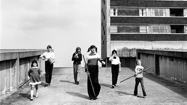 Children playing with a large tire on a rooftop concrete platform beside a brutalist apartment building.