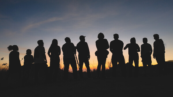 Silhouettes of approximately twelve people standing in a line against a golden sunset sky.