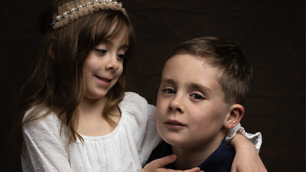 Young girl with tiara embracing boy from behind against dark background.