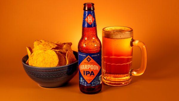 Bottle of IPA beer with glass mug and bowl of potato chips against warm orange background.