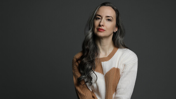 Woman with long dark hair wearing a cream and tan ribbed knit sweater against a gray background.