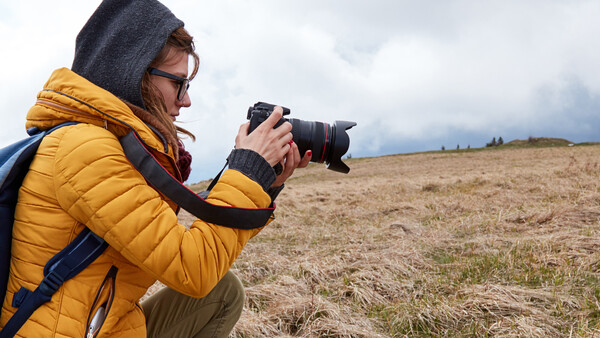 Photographer in yellow jacket and hood holding DSLR camera while crouching in open field.